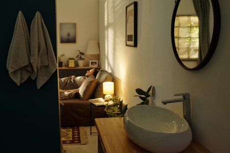 Caucasian man lying in bed appearing stressed with hand on forehead in softly lit bedroom, visible from bathroom with modern sink and towels hanging on wallの写真素材