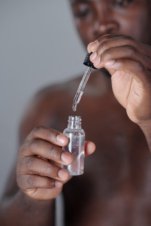 Vertical shot of Black man holding dropper bottle with clear liquid, gently squeezing contents into container, focus on hands and bottle, background blurred for emphasisの写真素材