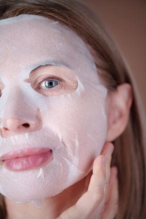 Vertical close-up portrait of woman with light skin wearing white moisturizing face mask while looking into camera, her hand gently touching face and background out of focusの写真素材