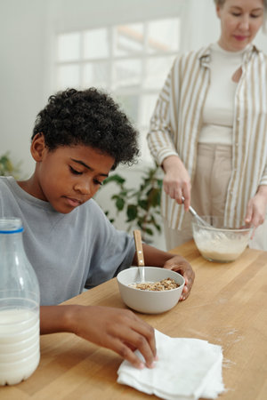 Young boy sitting at table eating cereal while adult standing in background with mixing bowl...
