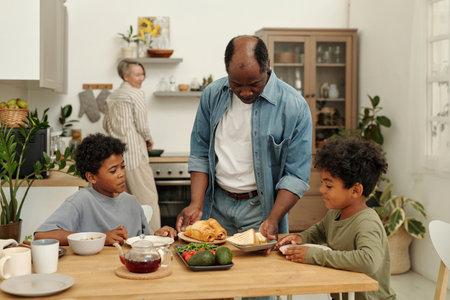 Diverse family sharing breakfast in cozy kitchen, with elder preparing food and children engaged in conversation. Scene captures warm family dynamics and cheerful atmosphereの写真素材