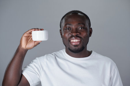 Portrait of smiling African American man holding small cream jar in studio setting smiling at camera with an expression of confidence and happinessの写真素材