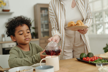Young boy pouring tea into cup while seated at kitchen table with breakfast spread of pastries, vegetables, and cereal bowl. Visible kitchen appliances and window in backgroundの写真素材