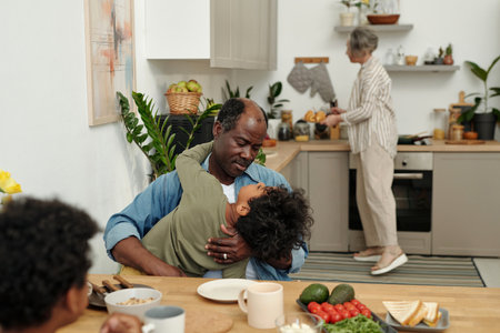 African American father holding child in bright kitchen while older woman prepares food in backgroundの写真素材
