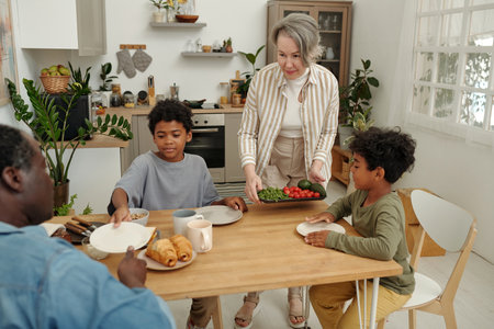 Caucasian mother bringing vegetables to African American children seated at kitchen table with food in modern, plant-filled kitchen interior decorated with shelves and windowsの写真素材