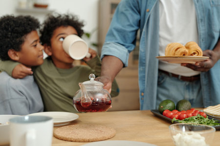 Man serving tea to children sitting at kitchen table with fresh pastries and vegetables visible creating a warm and inviting atmosphereの写真素材