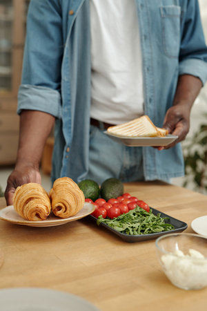 Person preparing healthy breakfast with fresh croissants, avocados, tomatoes, and greens. Table...