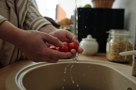 Hands of an elderly person washing fresh cherry tomatoes in kitchen sink with water running from faucet, surrounded by jars and containers on kitchen counterの写真素材
