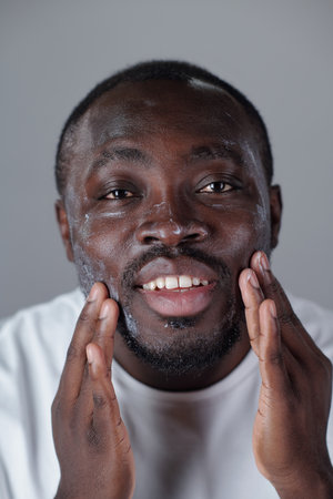 Portrait of smiling Black man enjoying skincare routine by applying cream on his face, displaying self-care and groomingの写真素材