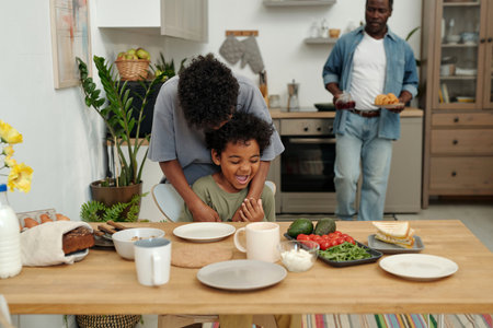 Family gathering in modern kitchen with mother hugging child and father holding plate of food. Various fresh ingredients and dishes displayed on wooden tableの写真素材