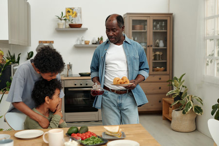 Smiling African American man holding jar of juice and plate with food while woman hugging child in cozy kitchen setting with plants, shelves, and wooden cabinetsの写真素材