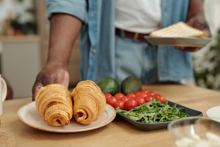 Close-up of hand serving croissants on plate with fresh avocados, cherry tomatoes, and leafy greens in background on kitchen counterの写真素材