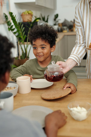 Smiling boy seated at kitchen table while drink being poured. Scene includes various bowls, plate, and green plants in background creating cozy settingの写真素材