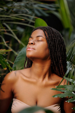 Young woman with closed eyes, enjoying sunlight while surrounded by tropical foliage. Relaxing...