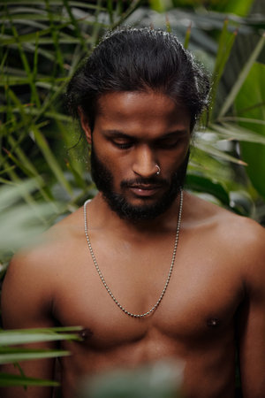 Man with beard and necklace standing in dense tropical rainforest, surrounding lush greenery present, eyes downcast, peaceful and contemplative expression, dark hair tied backの写真素材