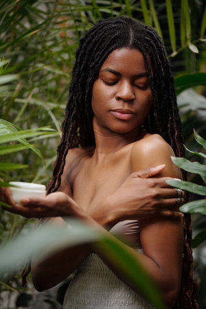 African American woman holding cream jar standing with closed eyes, embracing herself among lush green foliage, exuding calm and tranquility, embodying natural serenityの写真素材