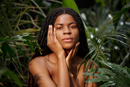 Portrait of Black woman touching face with hand while surrounded by lush green leaves, capturing serene and peaceful expression in a natural environmentの写真素材