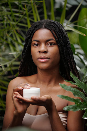 Portrait of young Black woman holding jar of skin cream in her hands, standing among lush green leaves, looking straight at camera, showcasing natural beauty and skincareの写真素材