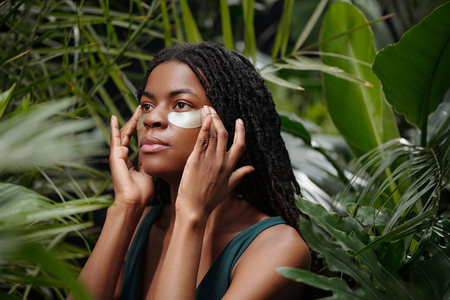 Woman with dark skin applying patches under eyes while sitting among lush green plants, with fingers lightly touching her faceの写真素材