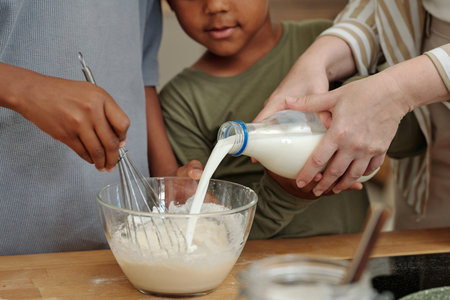 Close-up of hands mixing batter with boy watching attentively, with milk being poured into bowl....