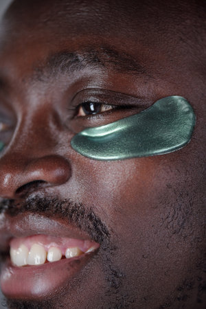 Vertical shot of smiling man with under-eye patches visible, highlighting skincare routine and self-care. Patches are green, covering area under his eyesの写真素材