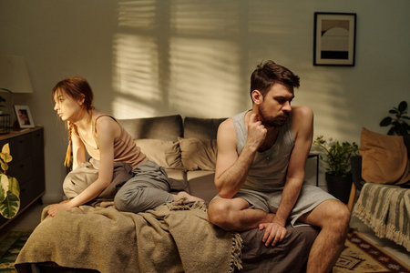 Man sitting looking down touching neck while young adult woman sitting on edge of bed looking away both appearing distant during family conflictの写真素材