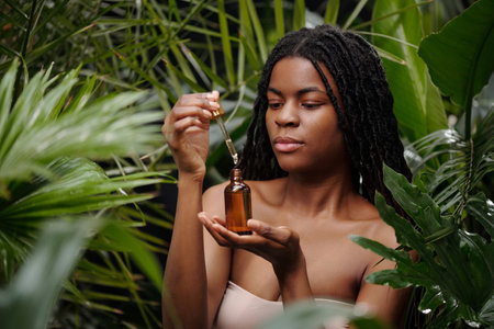 Woman with braided hair carefully holding amber bottle with dropper, surrounded by lush green plants, exuding calm and focus, with natural elements highlighting serene atmosphereの写真素材
