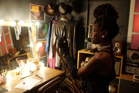Black woman young adult holding saxophone standing in dressing room preparing for jazz performance, musical instruments and stage costumes visible in background, looking away from camera