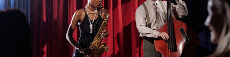 Black young adult woman playing saxophone and Caucasian middle aged man playing double bass performing on stage with red curtain background during jazz concertの写真素材
