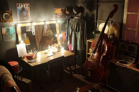 Musicians dressing room featuring upright bass, vintage reel-to-reel tape recorder, illuminated vanity mirror, jazz posters on wall, stage costumes hanging on rackの写真素材