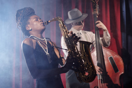 Black young adult woman playing saxophone with eyes closed, Caucasian middle aged man wearing hat playing double bass in background on stage with red curtain