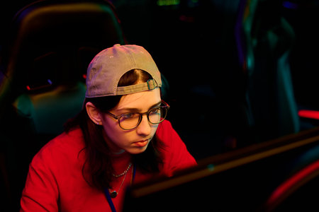 Caucasian teenage girl wearing glasses sitting at gaming station focusing on computer monitor...