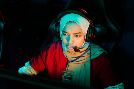 Portrait of young adult Muslim woman wearing headset participating in esports gaming competition, sitting at computer station, focused on screen, engaging in team gameplayの写真素材
