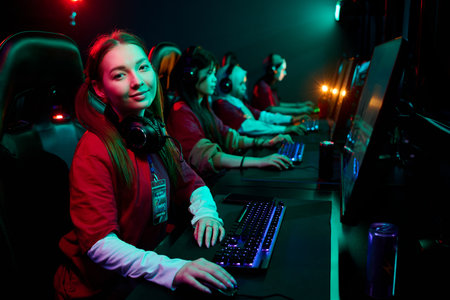 Teenage girl gamer sitting at computer desk wearing headset looking at camera with multiethnic teenage girls gaming team focused on monitors in e-sports competitionの写真素材