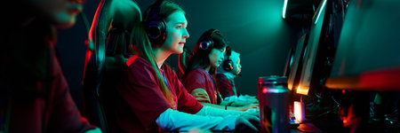 Group of multiethnic teenage girls wearing headsets competing in esports tournament, sitting side by side at gaming computers, focused on screens, illuminated by colorful lightsの写真素材