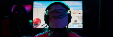 Teenage girl wearing headphones sitting in front of large gaming monitor playing video game in dark...
