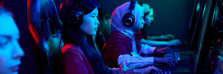Diverse group of teenage girls sitting at gaming computers wearing headsets, focusing on playing...