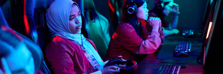 Multiethnic team of teenage girls sitting at gaming desks competing in esports tournament, wearing...