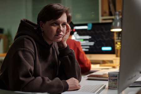 Caucasian young adult woman sitting at desk using computer, resting chin on hand, looking at monitor with scam call center interface visible on screenの写真素材