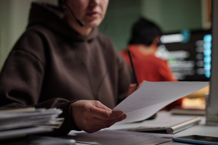 Young adult Caucasian woman wearing headset holding documents while working at computer in scam call center with blurred colleague in backgroundの写真素材
