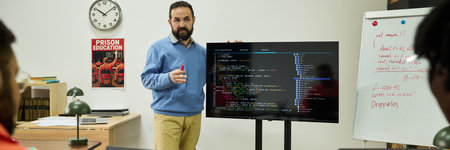 Middle aged Caucasian man teaching programming to group of students, standing beside large monitor displaying code, gesturing with marker, classroom setting with educational materialsの写真素材