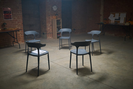 Empty chairs arranged in circle in spacious room with exposed brick walls, suggesting group therapy or support meeting setting, notebook resting on one chair, no people presentの写真素材
