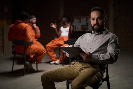 Caucasian middle aged man holding clipboard sitting in foreground, leading group therapy session with diverse young adults and teenagers in prison uniforms talking in backgroundの写真素材