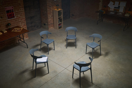 Empty circle of six modern chairs arranged in spacious brick room, single sheet of paper placed on one chair, suggesting group therapy or support meeting settingの写真素材