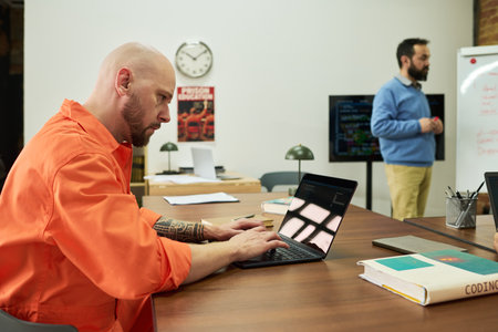 Caucasian man with shaved head and tattoos working on laptop at desk, Caucasian young adult man with...