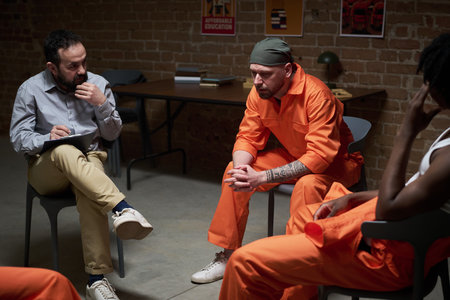 Caucasian man in orange prison uniform sitting with head down during group therapy session, with Black man and young adult Caucasian male counselor taking notes in correctional facilityの写真素材