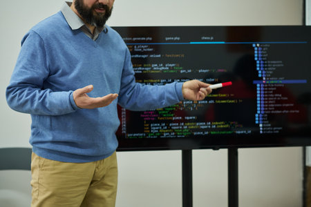 Middle aged Caucasian man teaching programming in prison classroom, holding marker and gesturing in front of large screen displaying code, focusing on educational instructionの写真素材