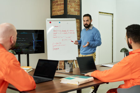 Middle aged Caucasian man teaching coding to two young adult men in orange prison uniforms, standing...