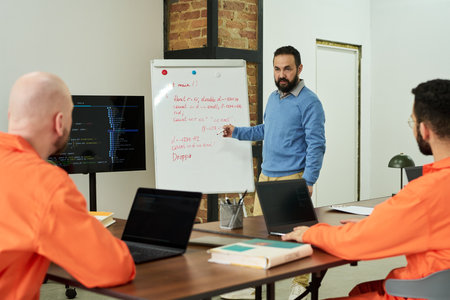 Middle aged Caucasian man teaching coding to twomen in orange prison uniforms, standing by...