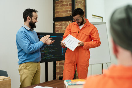 Middle aged man applauding young adult man wearing prison uniform receiving certificate in classroom...
